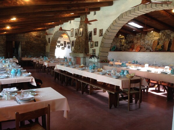 A long dining table set with white tablecloths and blue glassware, surrounded by stone walls and wooden beams. - Agriturismo Lu Branu (Arzachena)