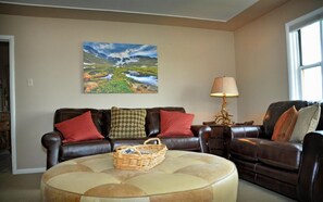 A living room with a brown leather sofa, a round ottoman, a lamp, and a painting of a landscape on the wall. - Monster Lake Ranch (Cody)