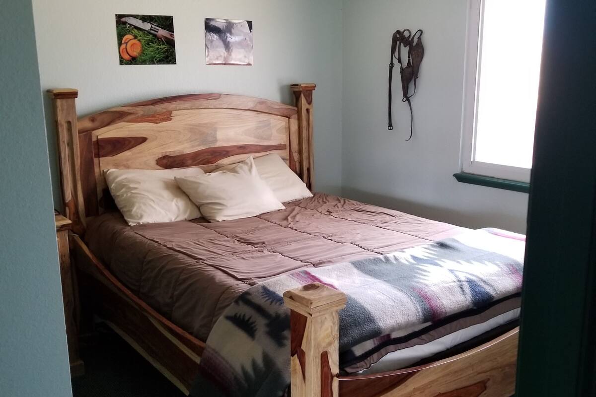 A wooden bed with a headboard, a window with blinds, and a wall with two framed pictures.