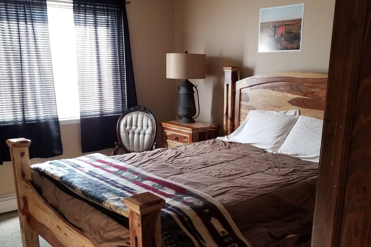 A bedroom with a wooden bed, a nightstand with a lamp, a chair, and a window with curtains.