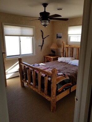 A bedroom with a wooden bed frame, a ceiling fan, a nightstand with a lamp, and a wall-mounted decoration. - Monster Lake Ranch (Cody)