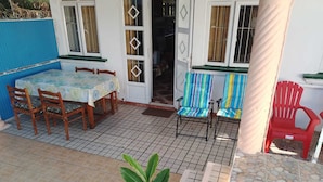 Uma área de pátio com piso de azulejos, uma mesa de madeira com cadeiras, uma cadeira colorida listrada e uma cadeira de plástico vermelha.