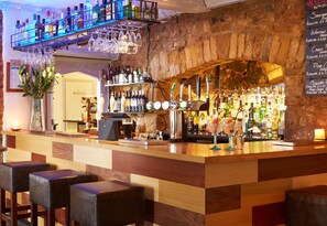 A bar area with a wooden counter, bar stools, a wine rack, and a stone wall. - Deddington Arms (Banbury)