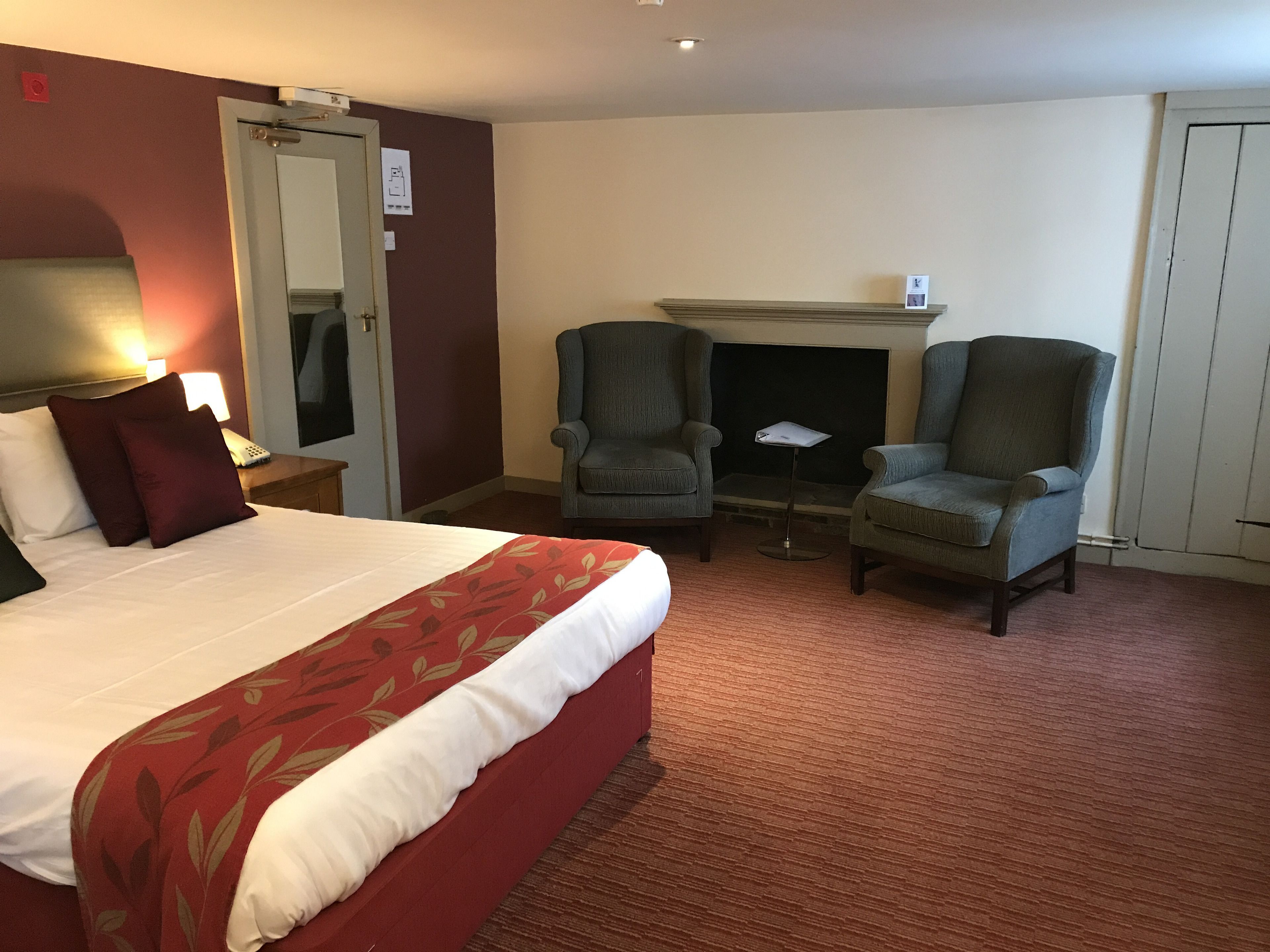 A hotel room with a bed, two armchairs, a small table, a fireplace, and a door with a sign.