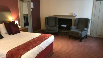 A hotel room with a bed, two armchairs, a small table, a fireplace, and a door with a sign.