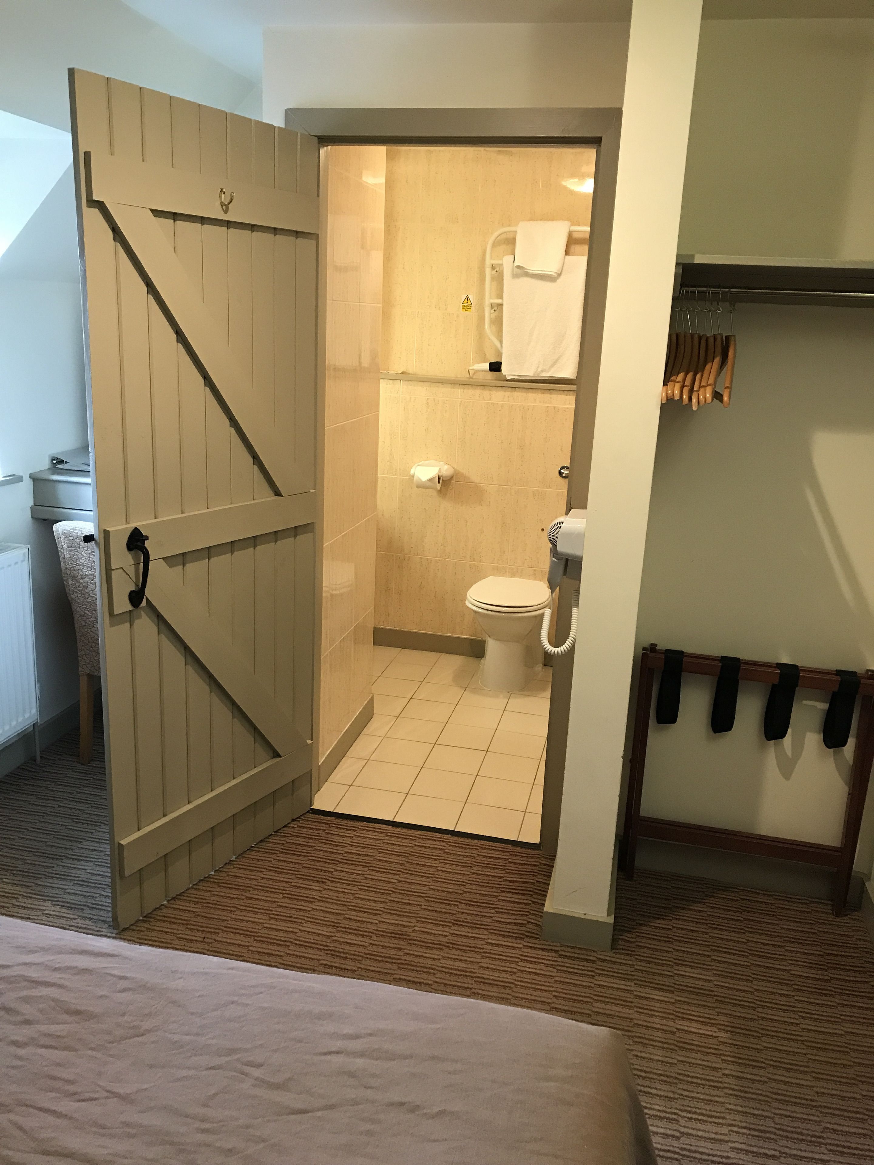 Quarto de hotel com porta de madeira que dá para um banheiro. O banheiro possui um vaso sanitário, um porta-toalhas e uma área de chuveiro.