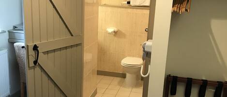 A hotel room with a wooden door leading to a bathroom. The bathroom has a toilet, a towel rack, and a shower area.