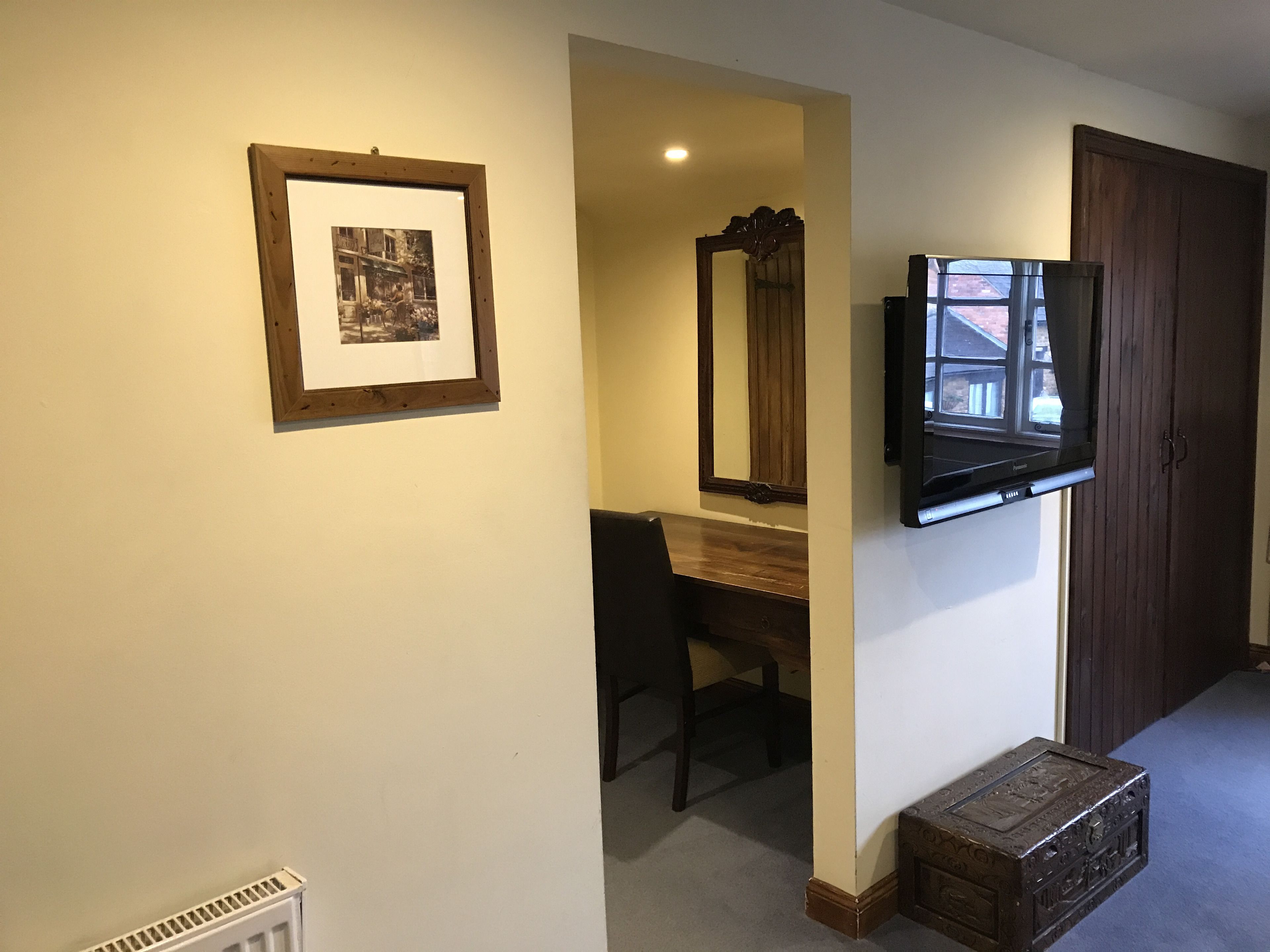 Un couloir avec un tableau encadré, un miroir, un téléviseur fixé au mur, un bureau en bois et une porte.