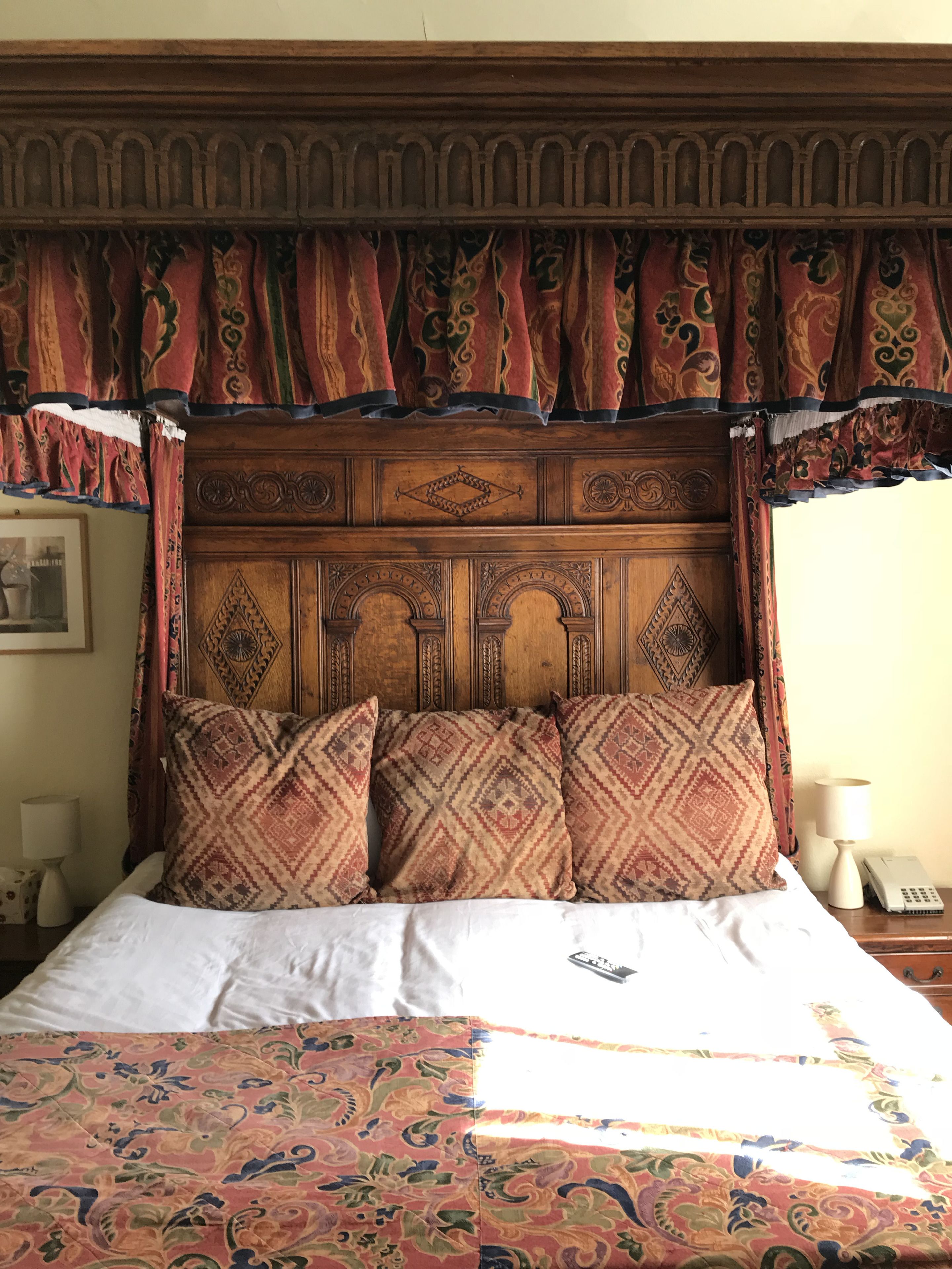 Uma cama com colcha estampada, cabeceira de madeira com esculturas detalhadas e saia decorativa.