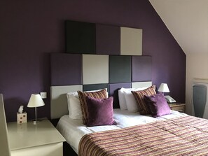 A hotel room with a modern design, featuring a large bed with a striped bedspread, two nightstands with lamps, and a unique geometric headboard. - Deddington Arms (Banbury)
