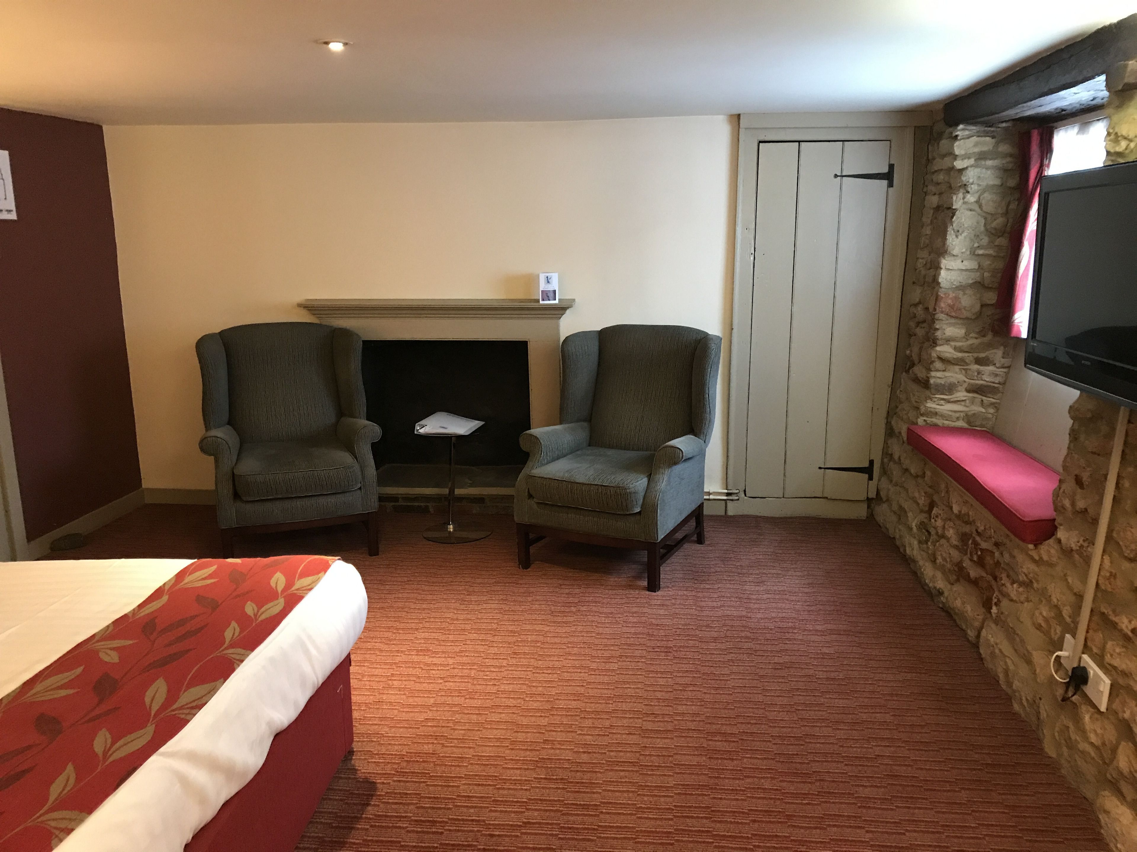 A hotel room with a bed, two armchairs, a small table, a fireplace, a stone wall, and a TV mounted on the wall.