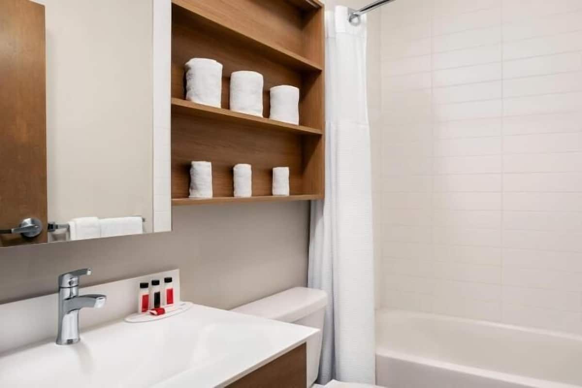 A bathroom with a white sink, a mirror, a shower with a glass door, and a shelf with rolled towels.