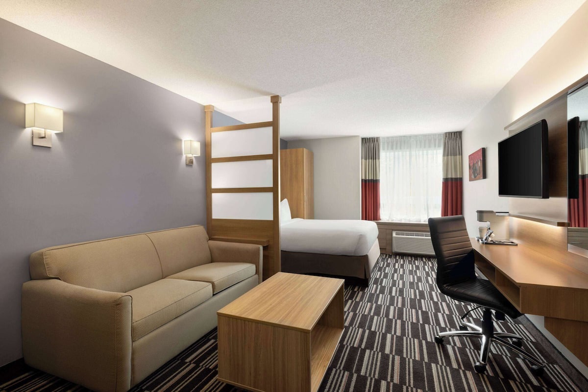 A hotel room with a sofa, a bed, a desk, a chair, a TV, and a window with curtains.