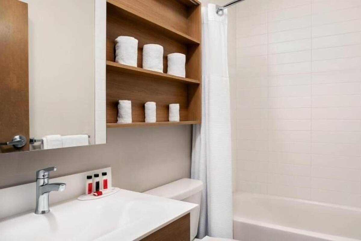 A bathroom with a white sink, a mirror, a shower with a glass door, and a shelf with rolled towels.
