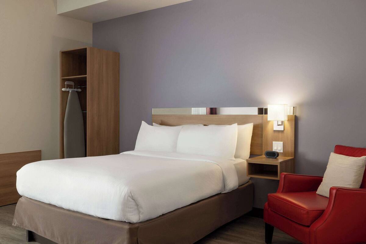 A hotel room with a large bed, a red armchair, a wooden wardrobe, and a bedside table with a lamp.