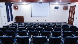 A conference room with rows of black chairs, a large white projection screen, wooden paneling, and a clock on the wall. - JS Park Hotel (Sao Lourenco)