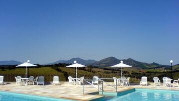 Piscina com cadeiras e guarda-sóis brancos, emoldurada por uma paisagem montanhosa.