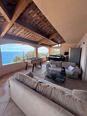 Un salon confortable avec un canapé, une table basse et une table de baby-foot. De grandes fenêtres offrent une vue sur la mer.