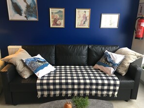 En svart lädersoffa med en rutig pläd, flera kuddar och inramade konstverk på en blå vägg.