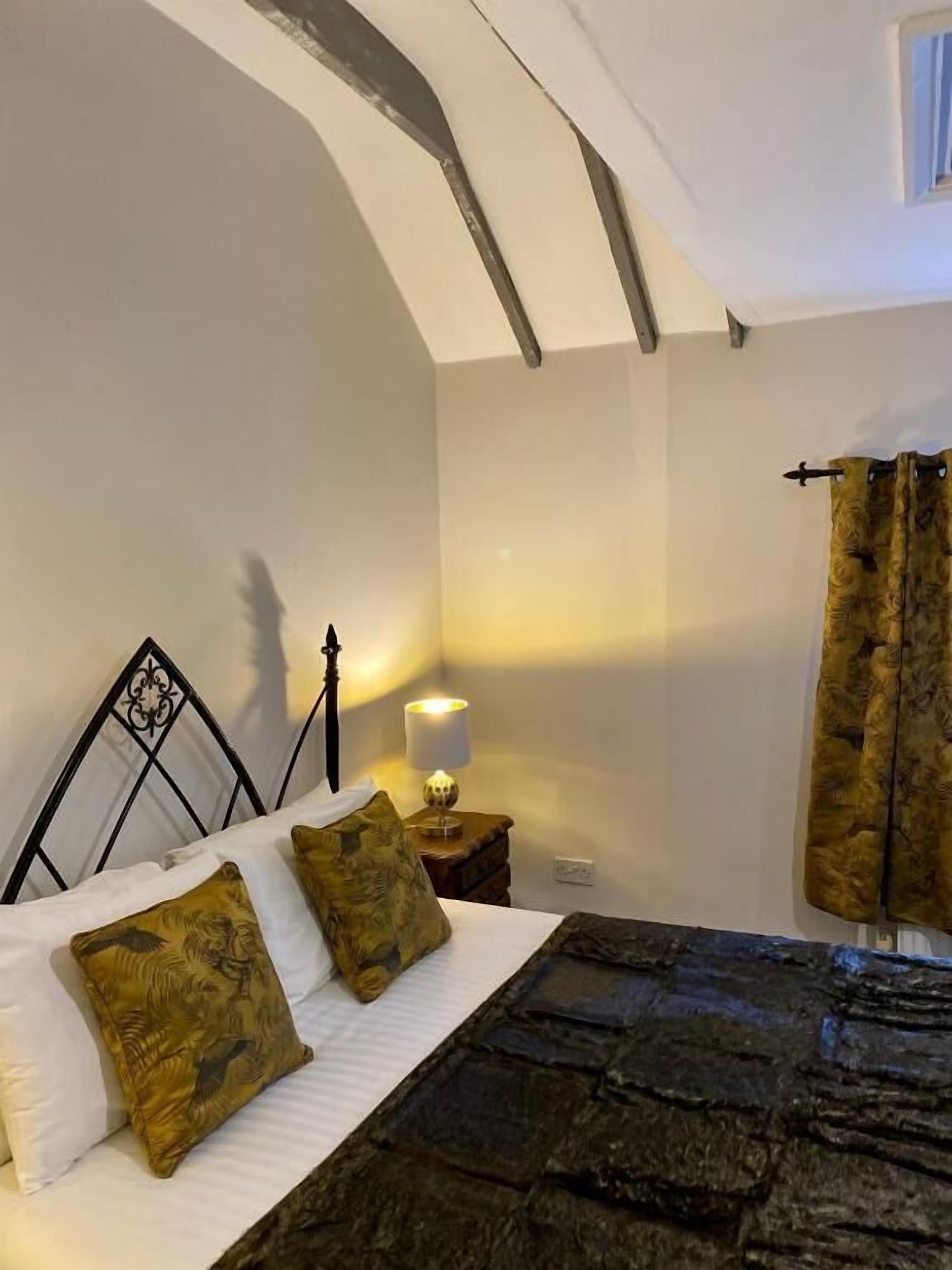 Un dormitorio con cabecera de piedra, cama con dos almohadas, mesita de noche con lámpara y ventana con cortinas.