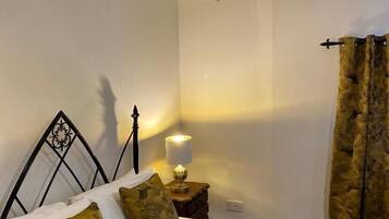 Un dormitorio con cabecera de piedra, cama con dos almohadas, mesita de noche con lámpara y ventana con cortinas.