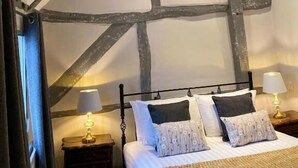 Quarto com vigas de madeira aparentes, cama com roupa de cama branca, duas luminárias de cabeceira e uma janela com cortinas.