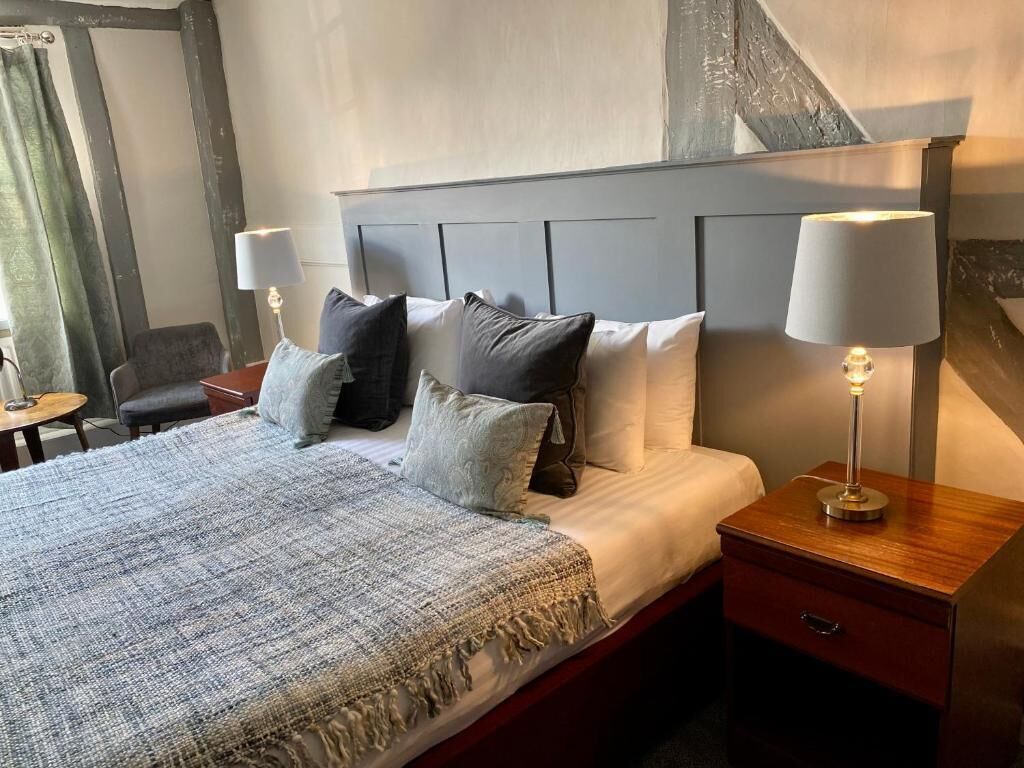 Una cama bien hecha con un edredón gris y almohadas, dos lámparas de noche y una mesita de noche de madera.