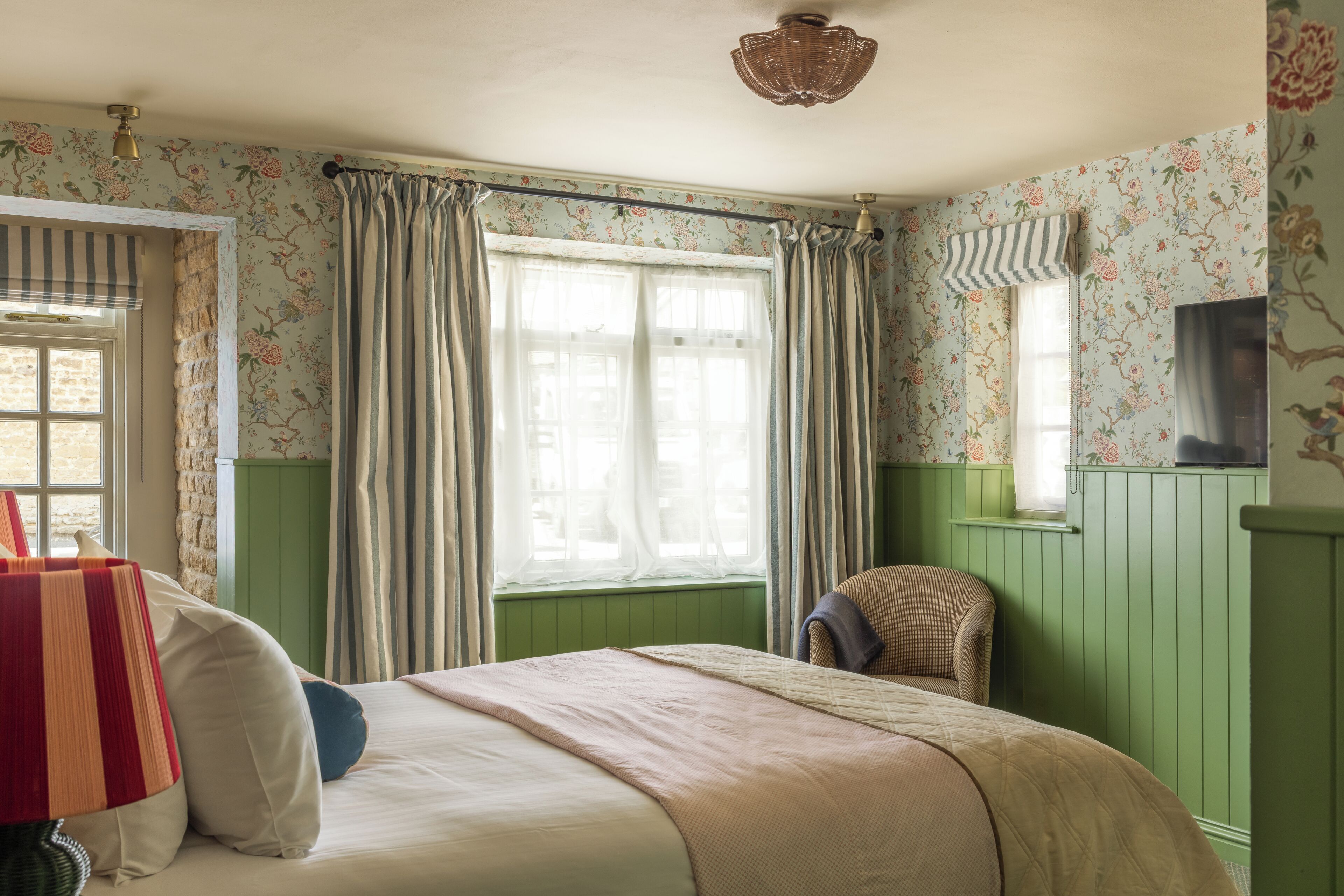 Una habitación con cama, una silla, un ventanal con cortinas y un papel tapiz floral en la pared.