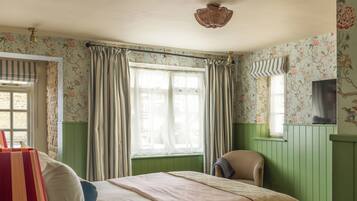 Una habitación con cama, una silla, un ventanal con cortinas y un papel tapiz floral en la pared.