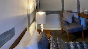 Quarto com vigas de madeira aparentes, uma cama com almofadas, uma cadeira, uma mesa com abajur e um tapete estampado.