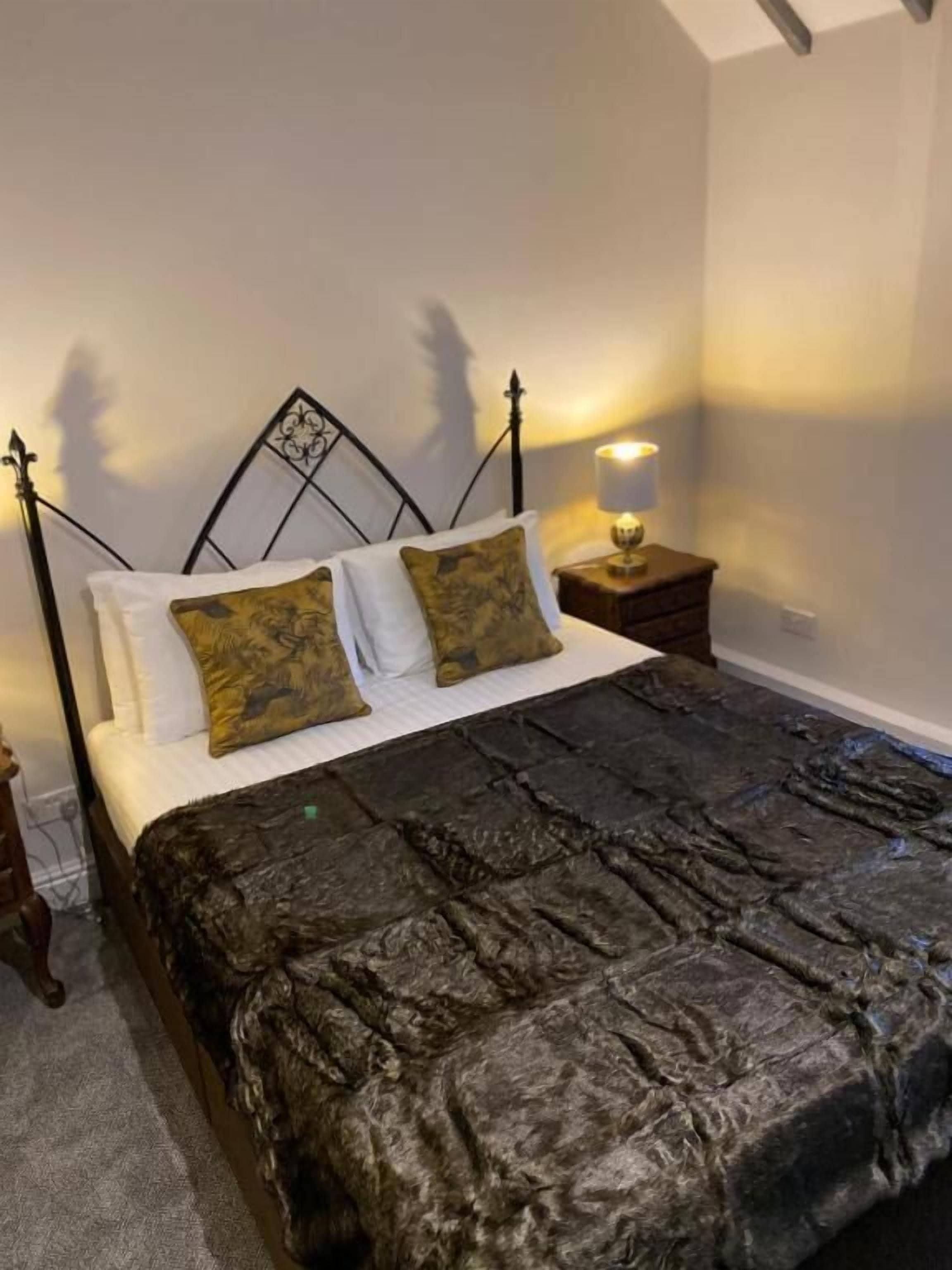 Ein Bett mit Metallgestell, zwei weiße Kissen und zwei gemusterte Kissen. Ein Nachttisch mit Lampe steht neben dem Bett.