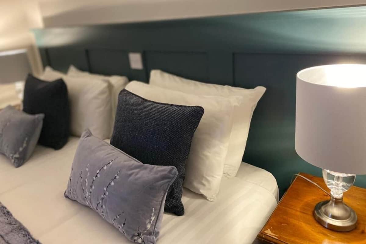 Una cama bien hecha con almohadas blancas y grises, un cabecero oscuro y una mesita de noche con una lámpara.