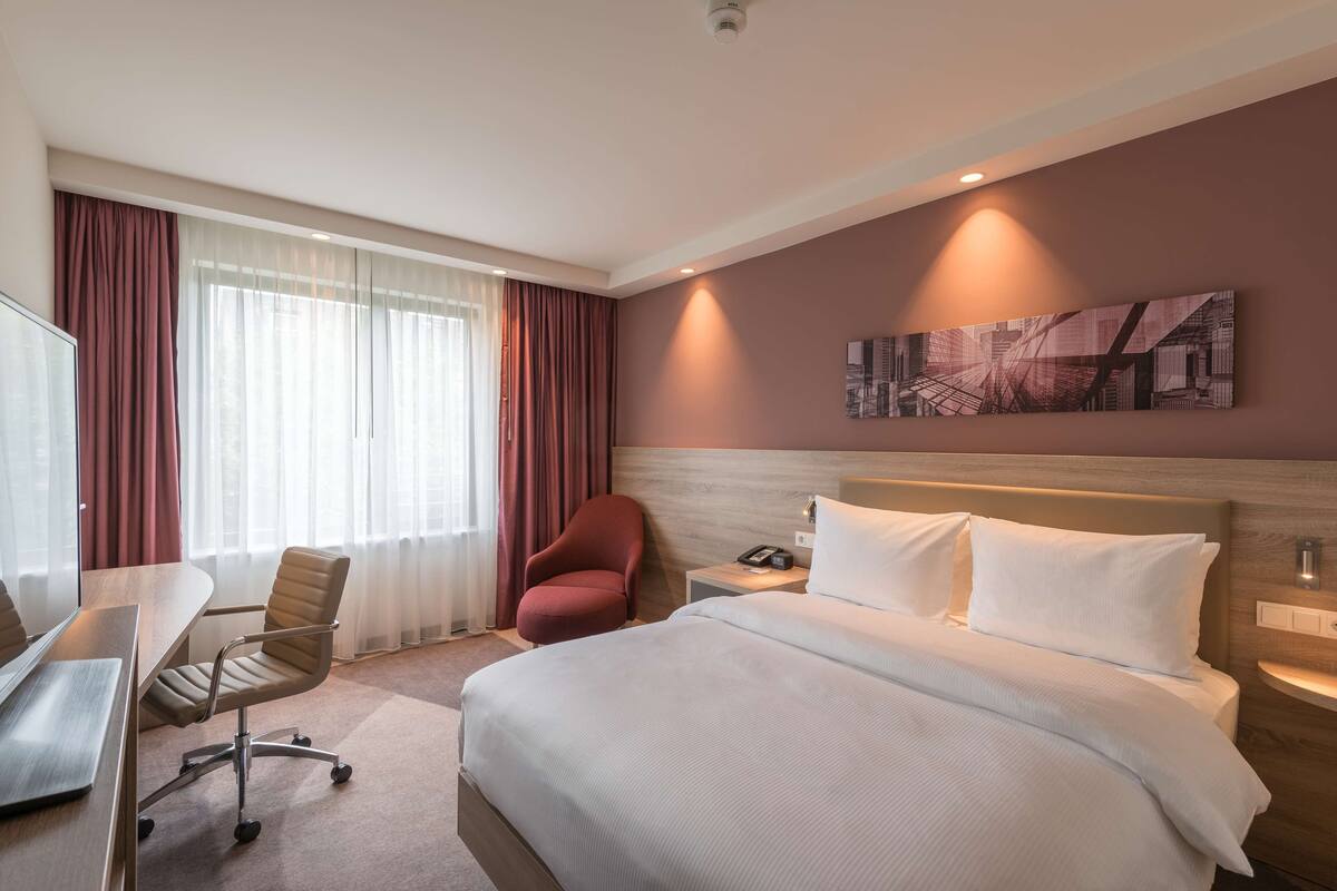 Ett hotellrum med en stor säng, ett skrivbord med en stol, en röd fåtölj och en tavla med en stadsbild på väggen.