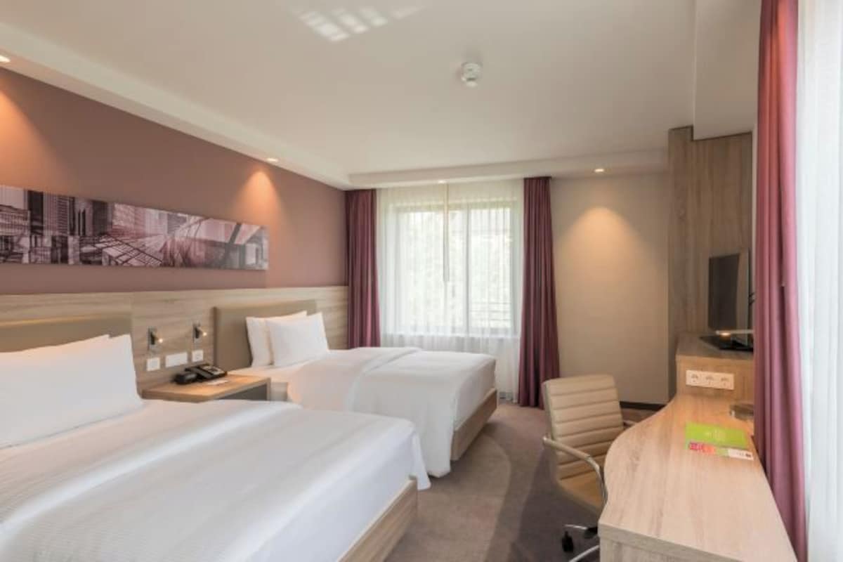 A hotel room with two beds, a desk, a chair, a TV, and a window with curtains.