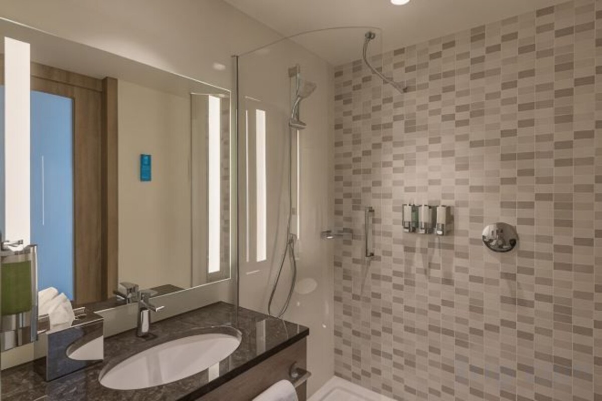 A modern bathroom with a large mirror, two sinks, a shower, and a tiled wall.