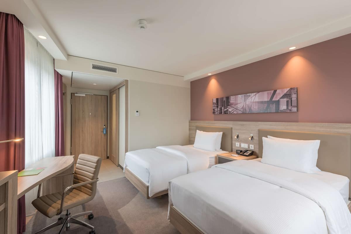 Ett hotellrum med två sängar, ett skrivbord och en stol. Det finns en tavla på väggen och en dörr som leder till ett annat rum.