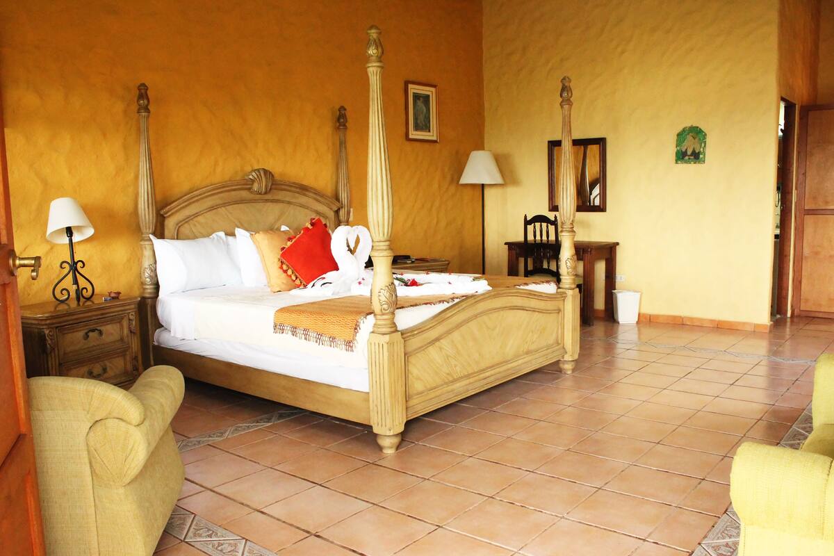 A four-poster bed with a wooden headboard, a bedside table with a lamp, and a chair in a room with tiled flooring and yellow walls.