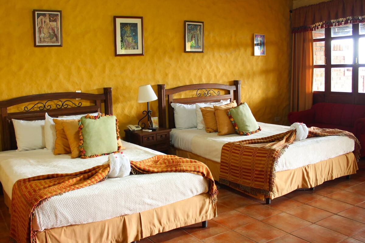 A hotel room with two beds, each with a wooden headboard, white linens, and multiple pillows. There are framed pictures on the wall, a nightstand with a lamp, and a window with curtains.