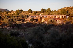 A modern house with a flat roof, large windows, and solar panels, surrounded by a dry, hilly landscape with sparse vegetation.