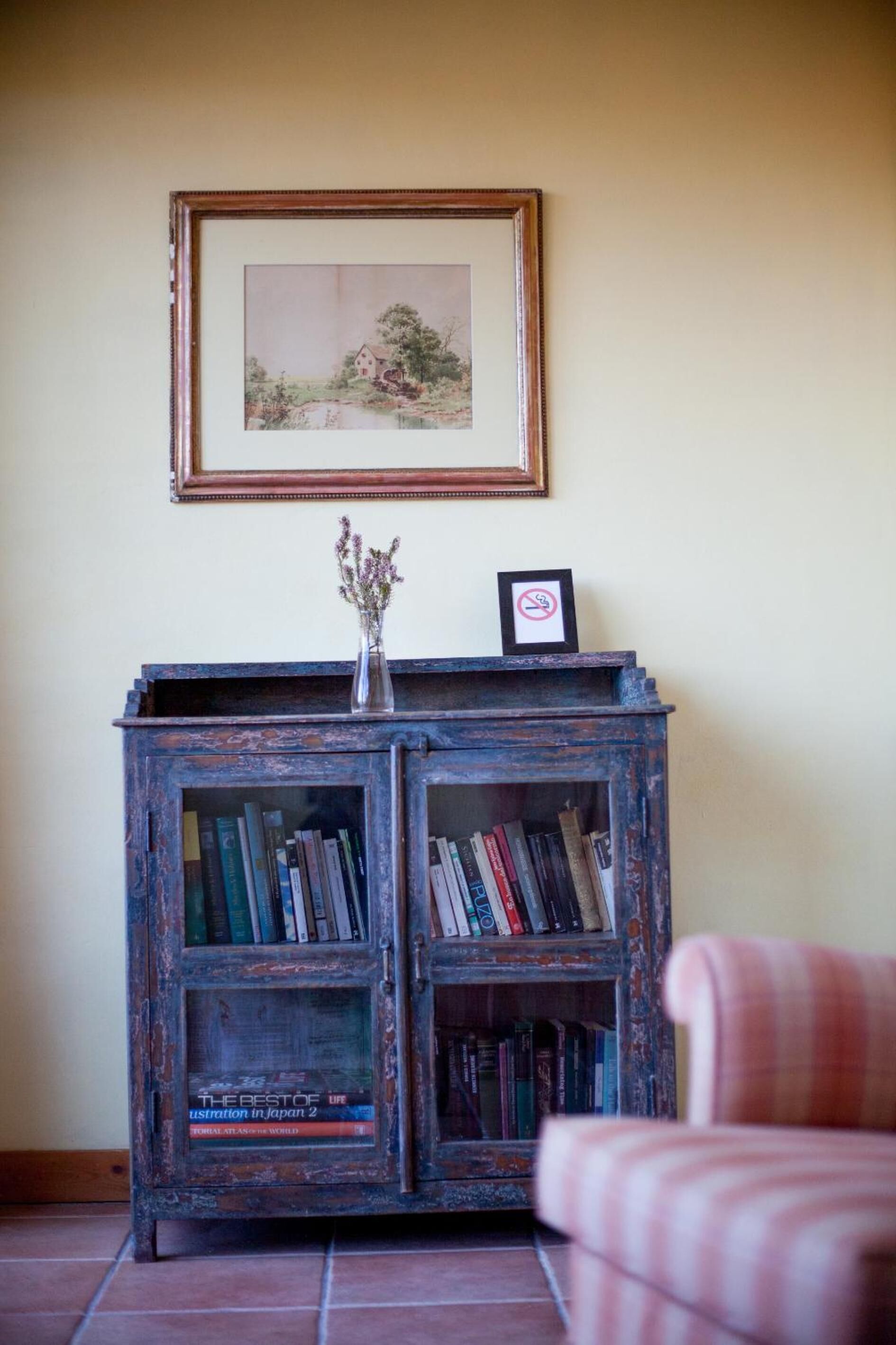 Uma estante de madeira rústica com livros e um vaso de flores, um quadro na parede e uma poltrona xadrez em primeiro plano.