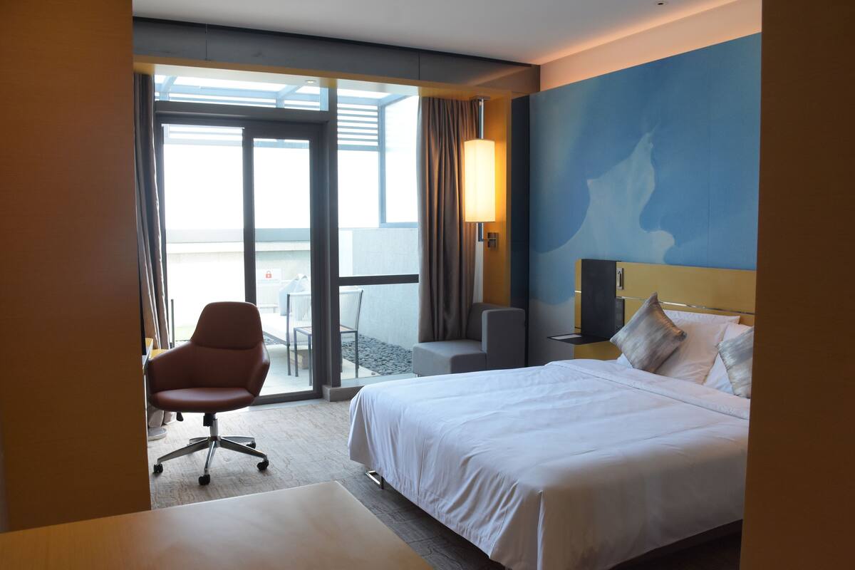 Habitación de hotel con cama, escritorio, silla, lámpara y vista al exterior.
