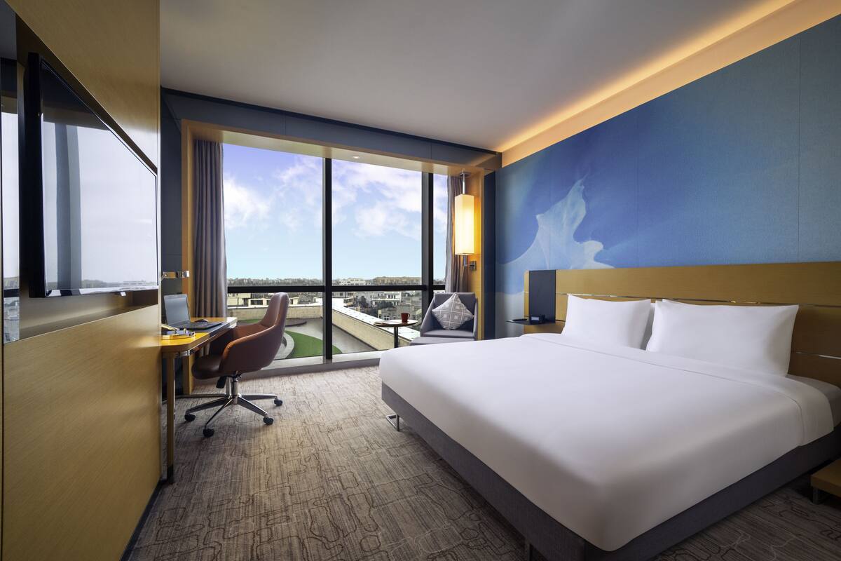 Habitación de hotel con una cama grande, un escritorio con una silla, vistas de la ciudad a través de amplias ventanas y un cuadro en la pared.