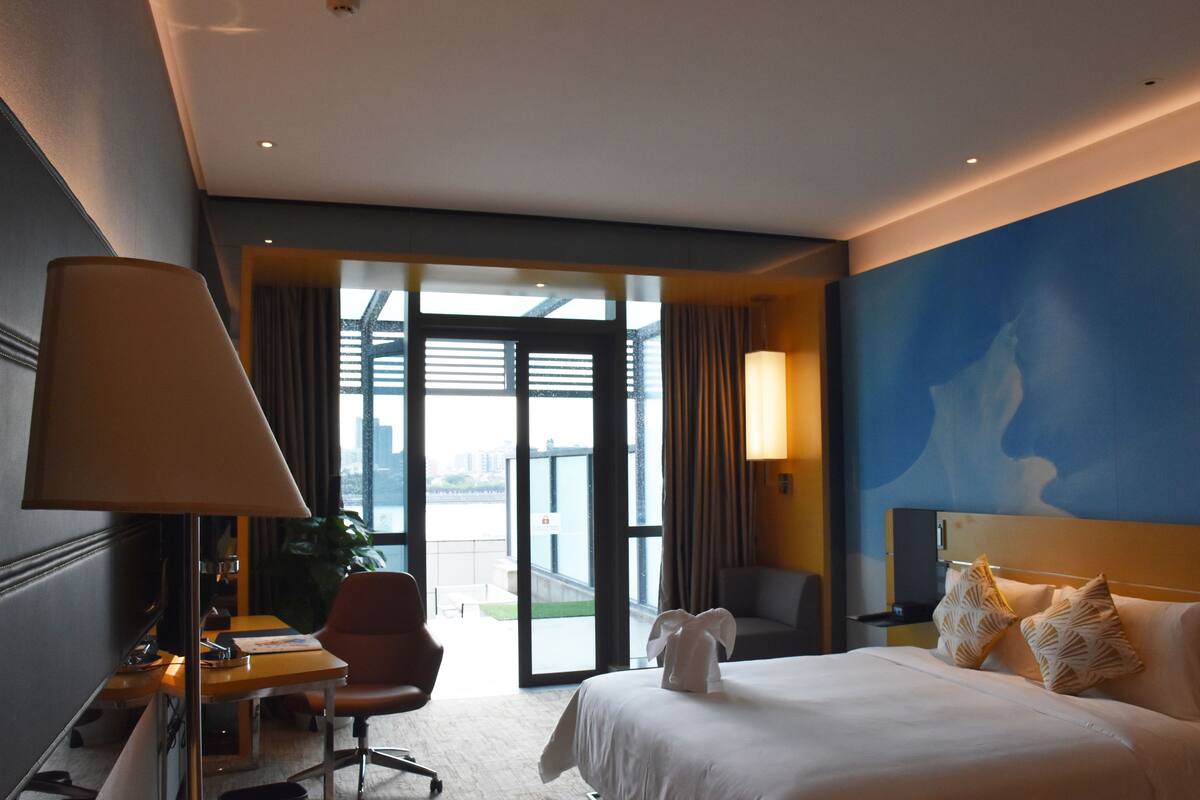 Habitación de hotel con cama, escritorio, silla, lámpara y vistas al exterior.