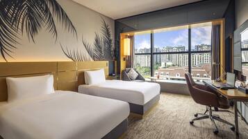 Habitación de hotel con dos camas, un escritorio con computadora, un ventanal con vista a la ciudad y una pared con un mural de hojas de palmera.