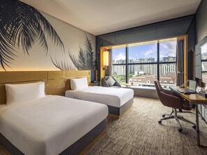 Habitación de hotel con dos camas, un escritorio con computadora, un ventanal con vista a la ciudad y una pared con un mural de hojas de palmera.