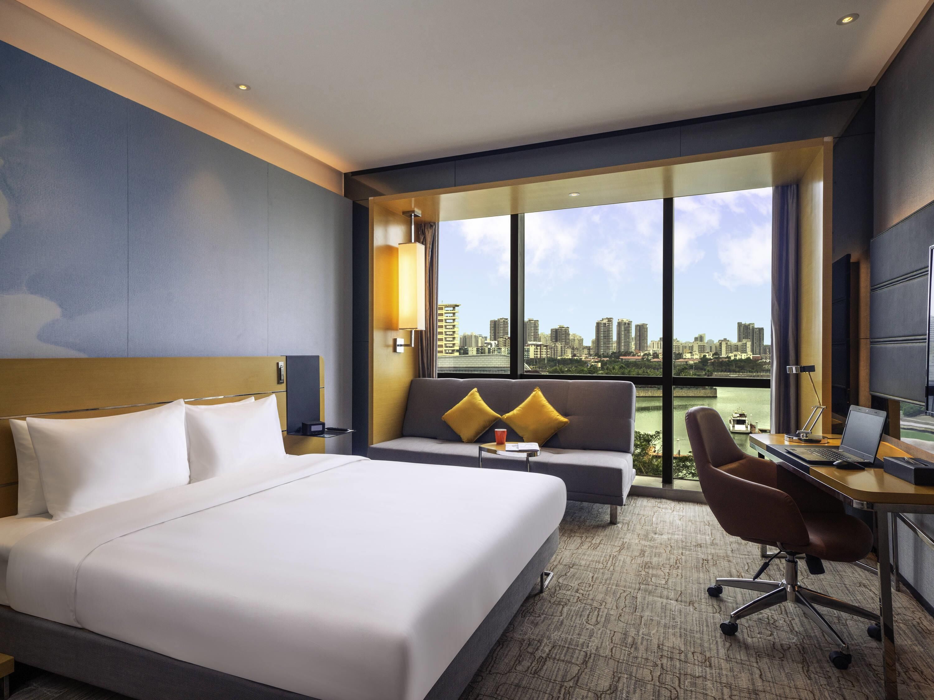 Une chambre d’hôtel avec un lit, un canapé, un bureau avec un ordinateur portable et une vue sur le paysage urbain.