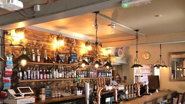 Zona de bar con mostrador de madera, taburetes y una variedad de botellas y vasos.
