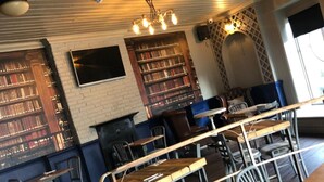 Un café acogedor con mesas y sillas de madera, una pared de ladrillo con una estantería y una lámpara decorativa en el techo.