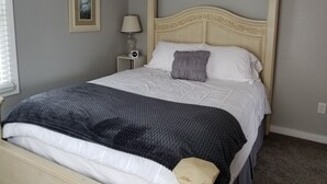 Ein ordentlich bezogenes Bett mit einer weißen und dunkelblauen Steude, einem grauen Kopfkissen und einem Nachttisch mit Lampe und Uhr.