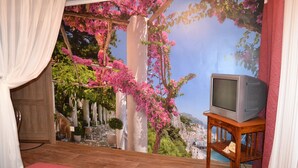 Une chambre à coucher avec un grand lit, un téléviseur posé sur un meuble en bois et une fresque murale représentant un paysage côtier.